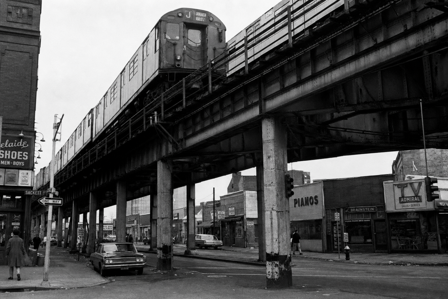 A New York City elevated subway train running above a Brooklyn street in the 1970s, shot from street level looking up